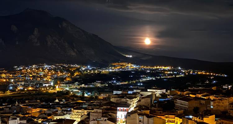 Nächtliches Panorama einer beleuchteten spanischen Stadt mit aufgehendem leuchtenden Mond hinter dunklen Bergen.
