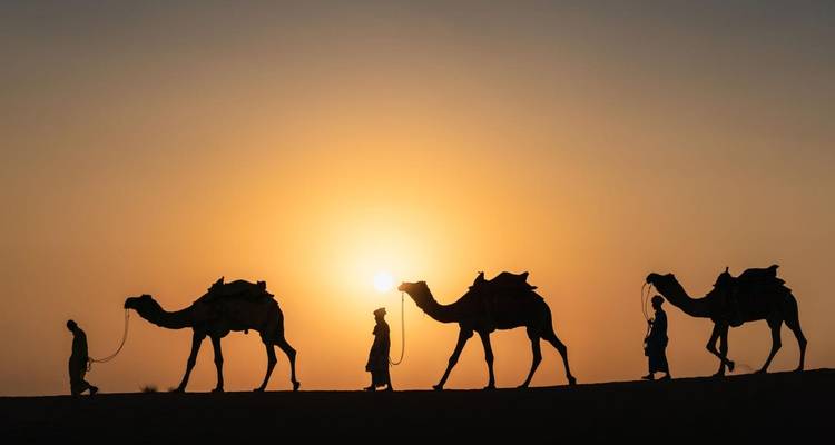Silhouettes de personnes conduisant des chameaux à travers les dunes du désert contre un coucher de soleil spectaculaire