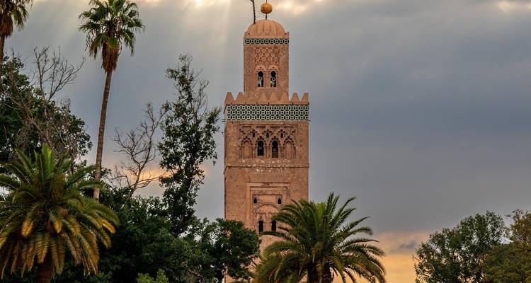 Minaret de la Mosquée Koutoubia encadré par des palmiers sous un ciel nuageux au crépuscule