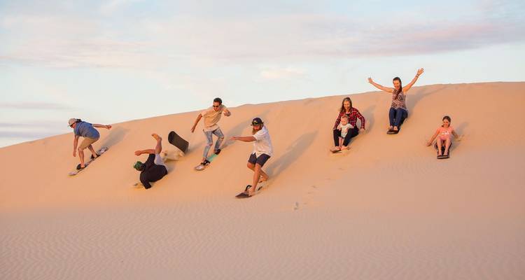 Groupe de voyageurs pratiquant le sandboard et glissant le long d'une haute dune dorée au coucher du soleil.