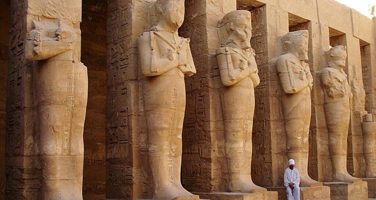 Massieve staande beelden staan in een zuilengang in de Karnak-tempel terwijl een bezoeker voorbijloopt.
