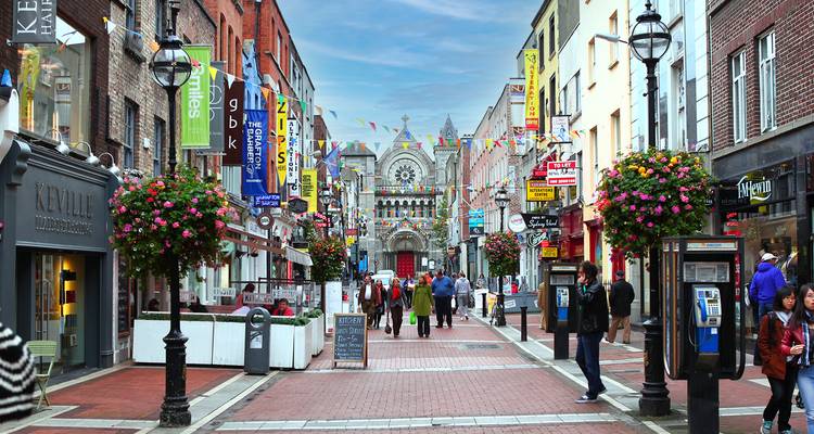 A vibrant pedestrian shopping street lined with flags, hanging flower baskets, and historic brick buildings.