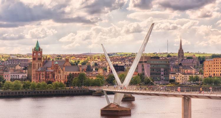 Modern footbridge with V-shaped supports spans a river toward a historic city with church spires and red-brick buildings.