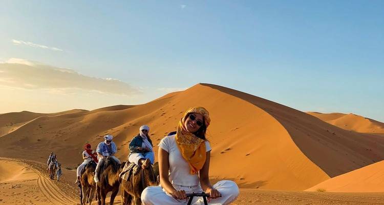 Une femme souriante est assise sur un chameau devant des dunes orange spectaculaires tandis qu'une caravane suit derrière.