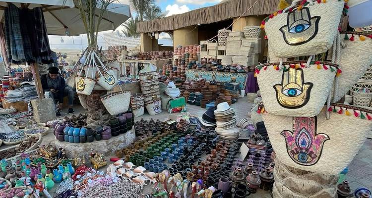 Un étal artisanal animé expose des rangées de poterie colorée et de sacs tissés avec le vendeur assis à proximité.