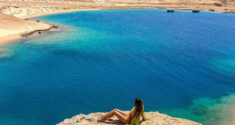 Woman relaxing on a rocky cliff edge overlooking vibrant turquoise waters of the Red Sea lagoon