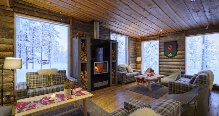 Warm lodge lounge with fireplace, stacked wood, and snowy forest visible through large windows.