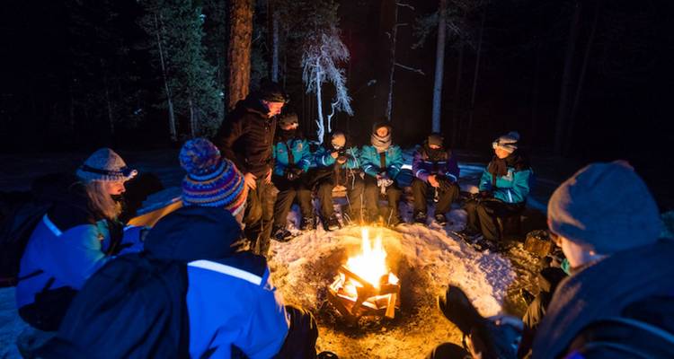 Group in winter gear gathered around a bright campfire on snowy ground at night.