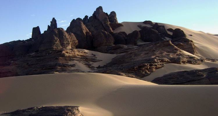 Dark jagged rock spires rising from sweeping pale sand dunes under clear sky.