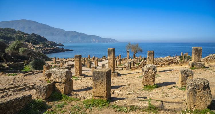 Ancient coastal Roman ruins with standing columns overlooking a brilliant blue sea and distant mountains.