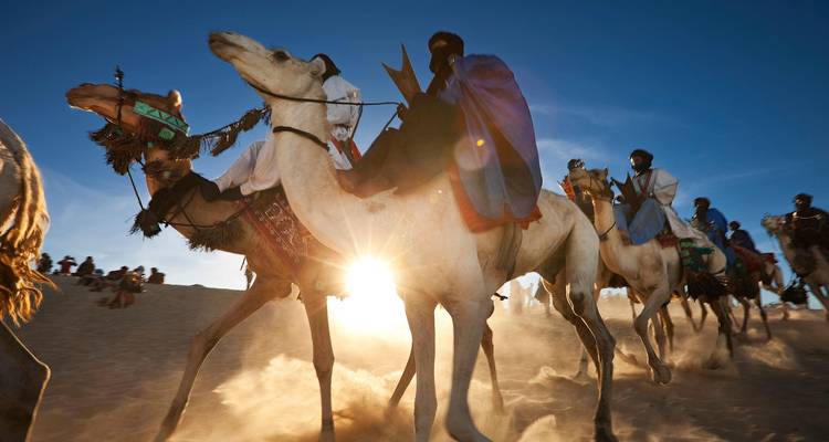 Camel riders charge through desert sand with a dramatic sunburst and flying dust