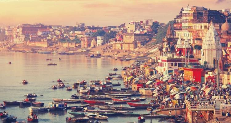 A bustling panorama of Varanasi’s ghats shows colorful buildings and boats lining the sacred Ganges River at dawn.