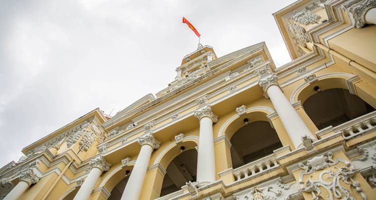 Upward view of the ornate Ho Chi Minh City Hall with columns and balconies beneath a cloudy sky
