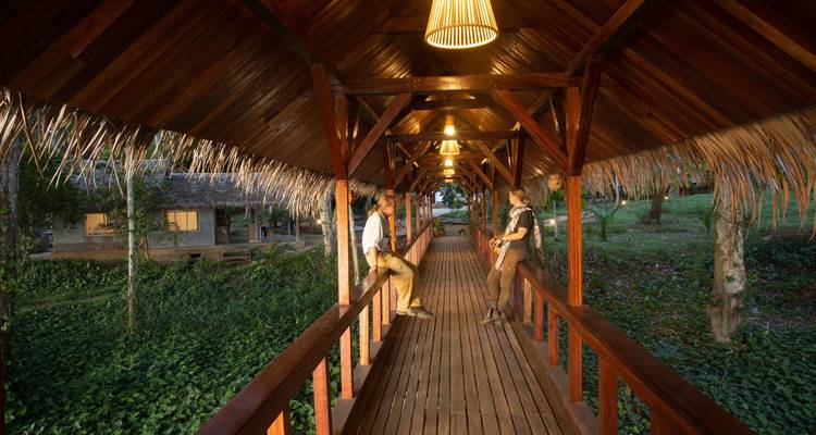 Two travelers chatting on a long covered wooden walkway lit by warm hanging lamps.