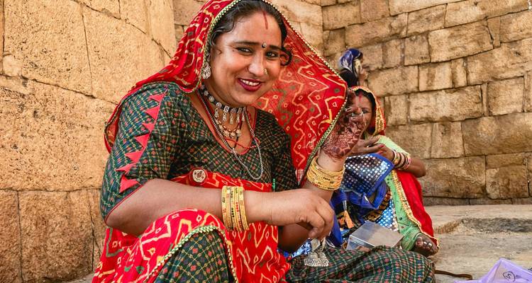 Lächelnde Rajasthan-Frau in einem leuchtend roten Sari zeigt aufwändige Henna-Verzierungen auf ihrer Hand vor einer Steinmauer.