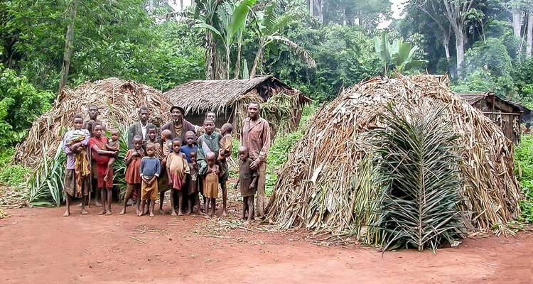 Community of indigenous villagers, adults and children, standing before dome-shaped leaf huts in a tropical forest clearing.