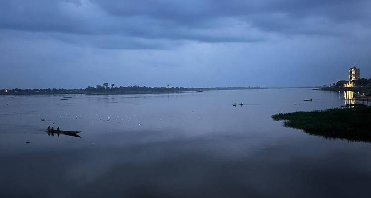 Wide evening view of a calm river under a moody blue sky with small wooden boats and distant shoreline lights.