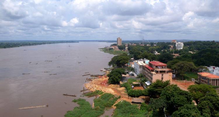 Aerial view along a wide river bordered by Bangui’s buildings and greenery under partly cloudy skies.