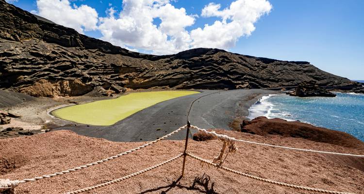 Beeindruckende grüne Lagune neben schwarzem Lava-Strand und erodierten Klippen unter blauem Himmel