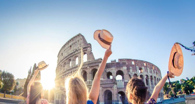 Group of young women waving hats toward the Colosseum in bright sunlight.