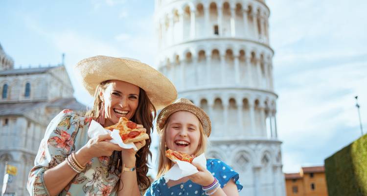 Mother and daughter eating pizza with the Leaning Tower of Pisa blurred behind.