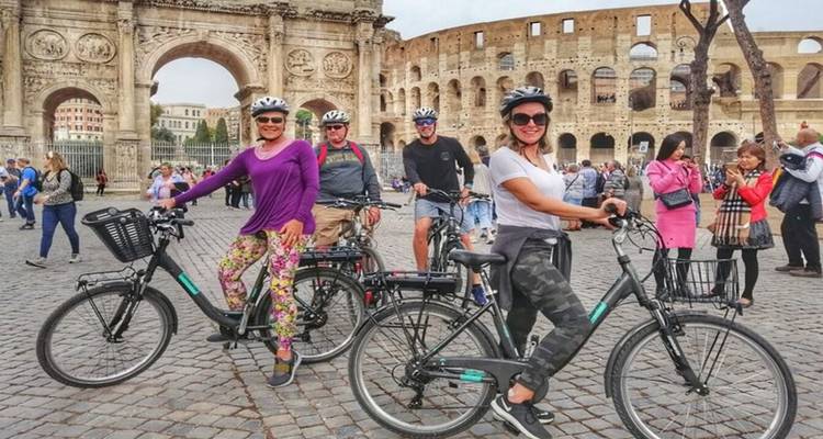 Cyclists pose with e-bikes in front of Rome’s Colosseum and Arch of Constantine on a cobbled plaza.