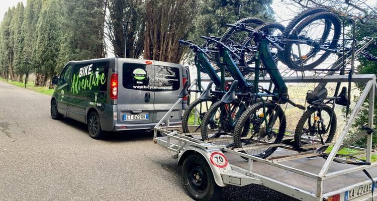 Van towing a trailer loaded with electric mountain bikes along a tree-lined country road.