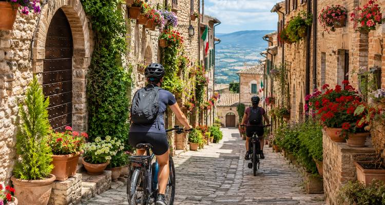 Cyclists ride through a picturesque stone alley adorned with flower pots in an Italian hill town.