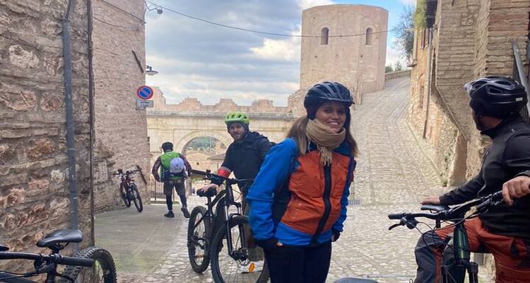 Group of cyclists stopping to chat in a stone alleyway beneath a medieval tower in Italy.