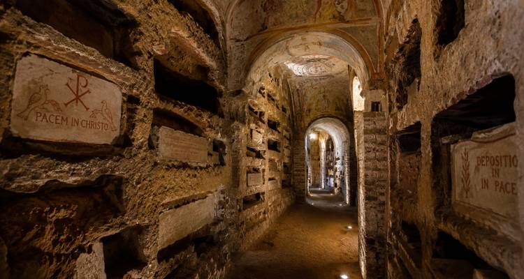 Illuminated corridor of ancient Roman catacombs with niches and faded Christian frescoes.