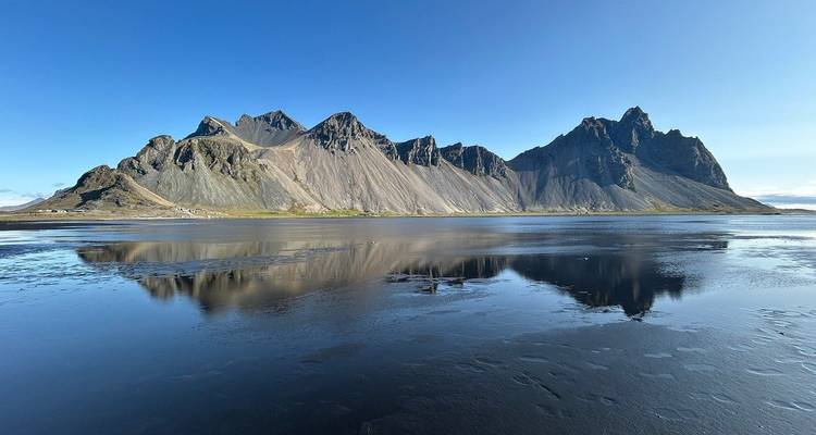 Mit Schnee bedeckte Gipfel des Vestrahorn, die sich in einer stillen Gezeitenlagune unter einem klaren blauen Himmel spiegeln.
