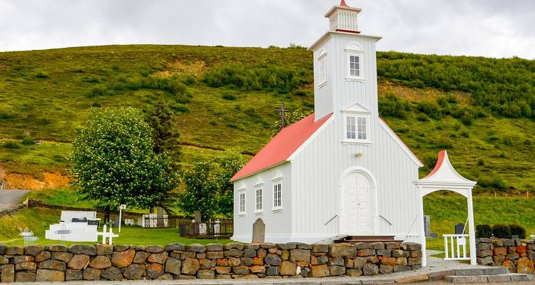 Charmante weiße Holzkirche mit rotem Dach, umgeben von einer Steinmauer und grünem Berghang.