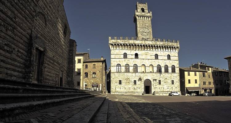 Palazzo médiéval en pierre avec une haute tour d'horloge dominant une place pavée vide sous un ciel bleu profond