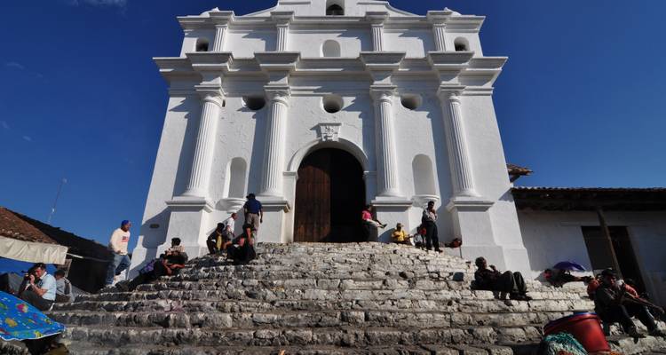 People sit on the stone steps of the white colonial church of Chichicastenango against a clear blue sky.