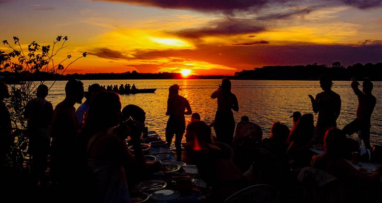 Silhouettierte Reisende genießen Abendessen und Getränke auf einer Flussufterasse, während eine lebendige orangefarbene Sonne über dem Amazonas untergeht.
