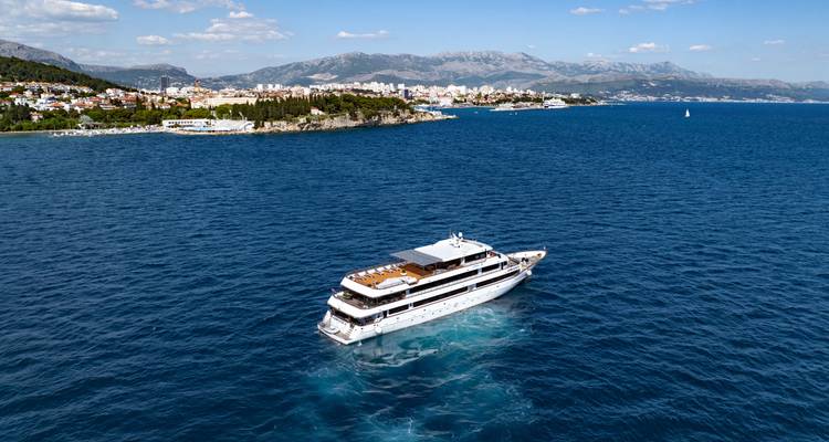 Drone view of cruise ship approaching a coastal city backed by mountains.