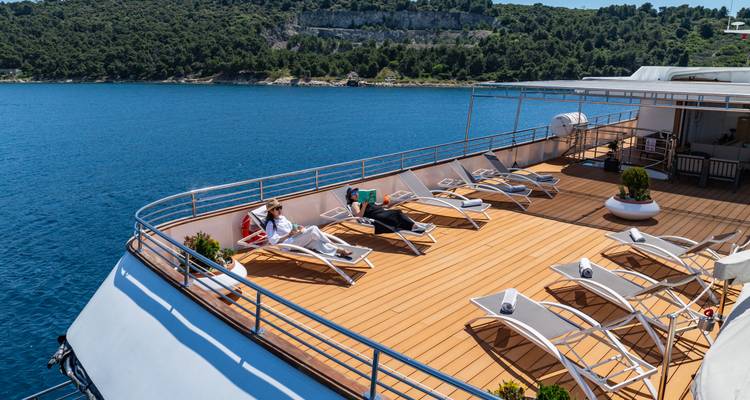 Two passengers reclining on sun loungers atop the yacht against bright blue water.