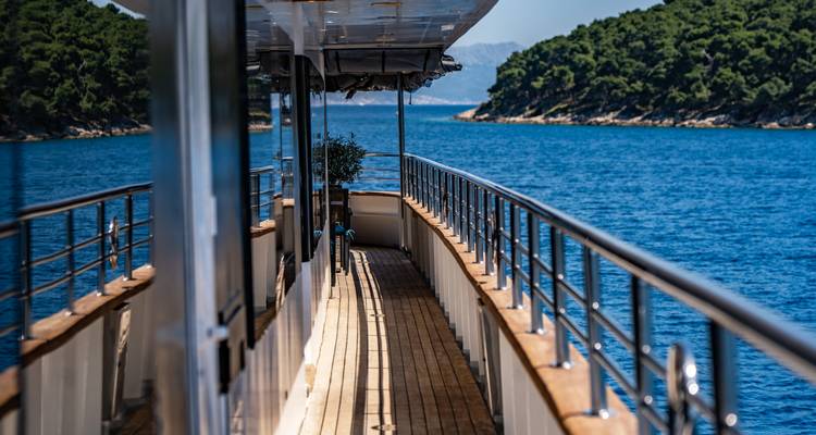 Yacht side deck walkway with view of tree-covered islands and clear blue water.