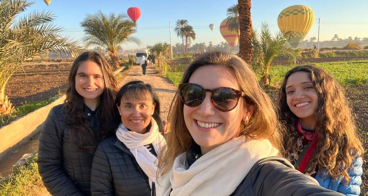 Four women smile for a selfie on a farm track while hot-air balloons rise behind them at sunrise.