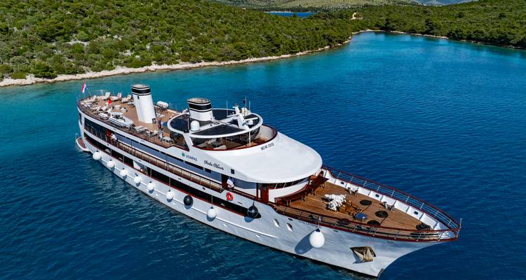 Close aerial angle of a sleek white cruise vessel navigating bright turquoise waters near a rocky coastline.