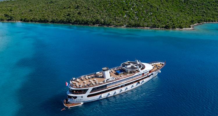 Luxury yacht anchored in a vivid blue inlet bordered by dense greenery, photographed from the air.