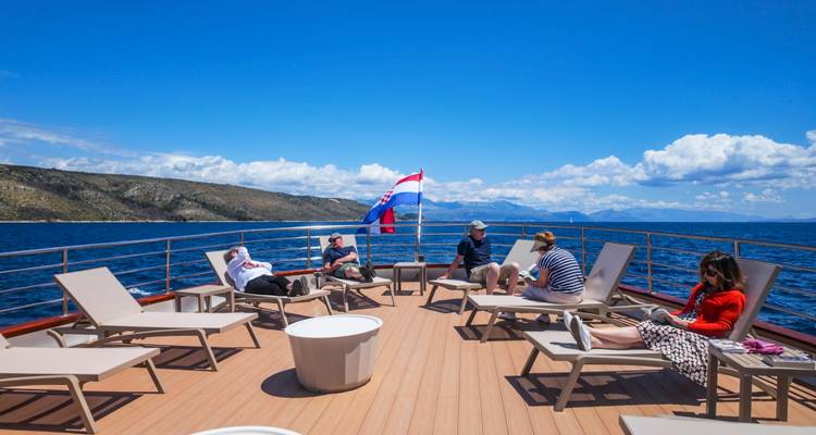 Passengers reclining on sun loungers atop a yacht deck under the Croatian flag with open sea views.