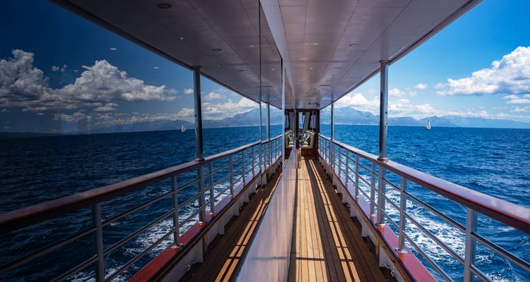 Symmetrical corridor deck view along a yacht with reflections of the sea and mountains on glass panels.