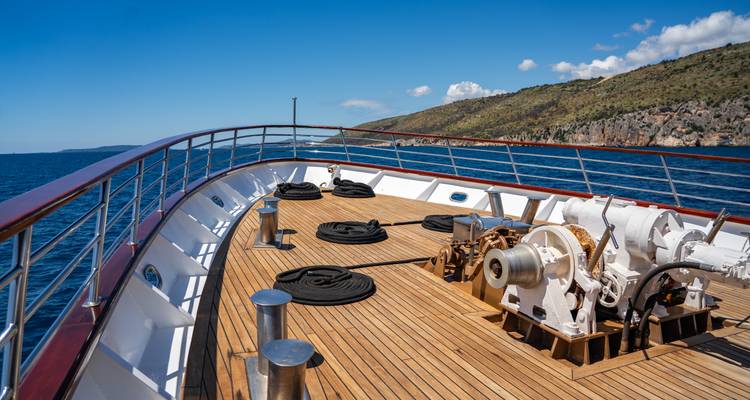 Forward deck of a yacht showing winches, ropes, and wooden decking against a blue sea backdrop.