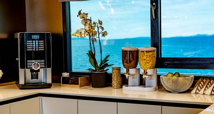 Modern self-serve coffee and snack station inside yacht with sea view through large window.