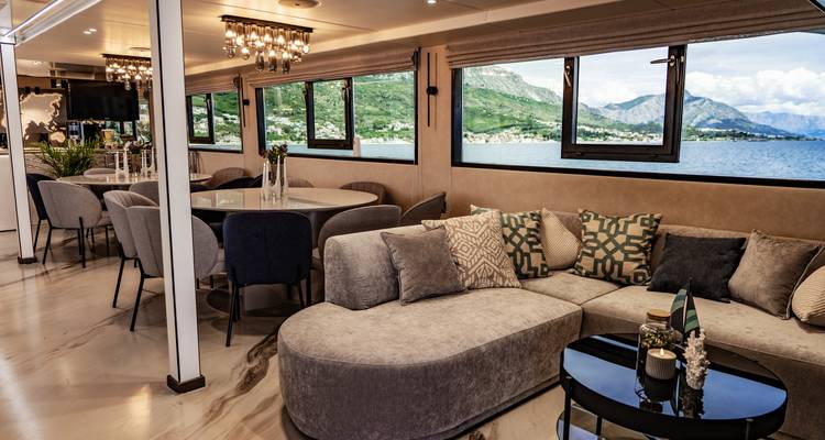Cozy yacht lounge with sofa, patterned cushions and sea-view windows overlooking hills.