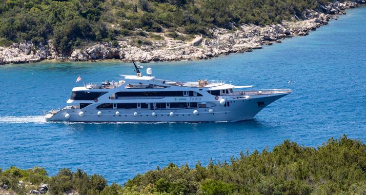 Full side view of luxury white yacht cruising turquoise waters near forested coast.