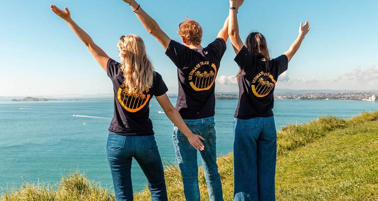 Three travellers in matching T-shirts raise their arms on a grassy clifftop overlooking turquoise coastal waters.