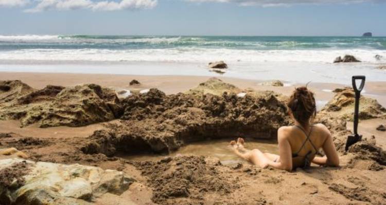 Traveller relaxing in a self-dug geothermal pool on a sandy beach with waves crashing nearby.