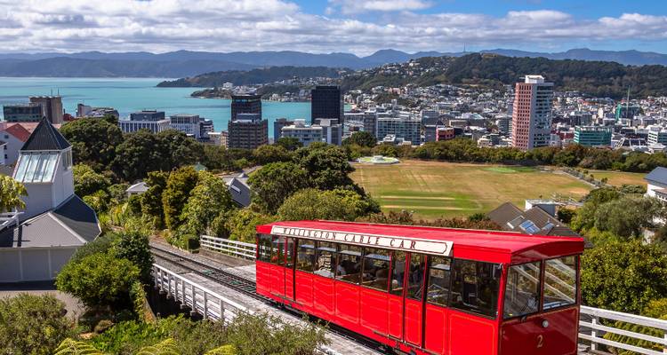 Iconic red Wellington Cable Car climbing a steep track with city skyline and harbour below.